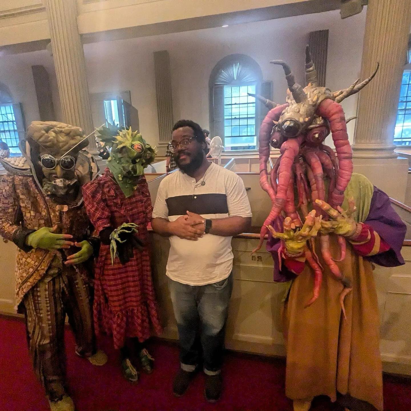 A dark-skinned man in glasses, a grey-and-black polo shirt, and jeans standing among a trio of masked, costumed persons, including one person wearing a pair of glasses with metal antennae emerging from the center, one person with tentacles emerging from its head, and another person with bug-like eyes and spikes emerging from its head.