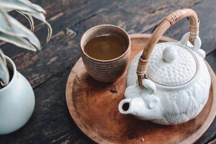 A white tea kettle and a brown cup full of tea, laying on a tray on a table beside a potted plant.